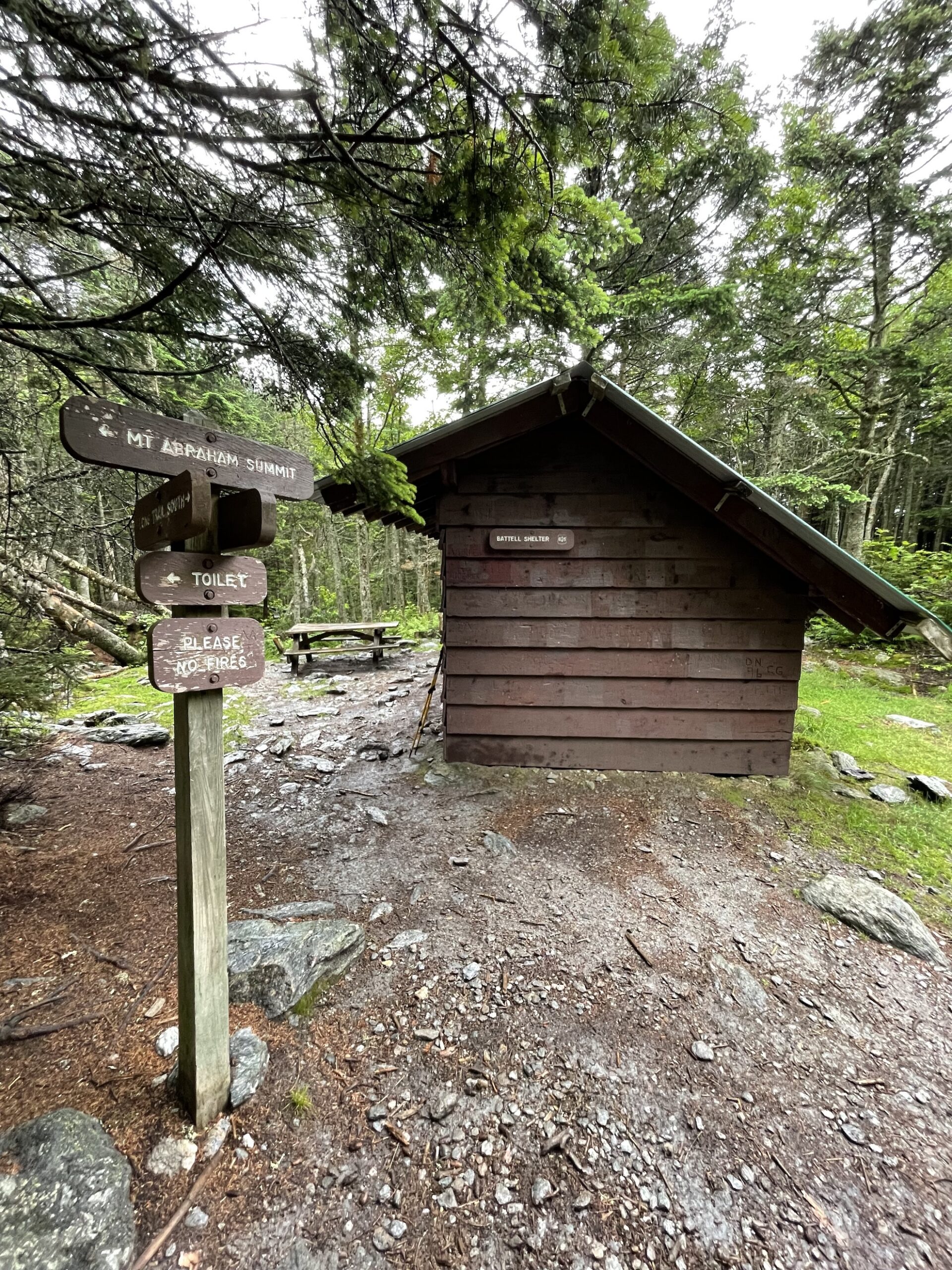 a trail sign sits in the foreground with a rustic leanto in the background