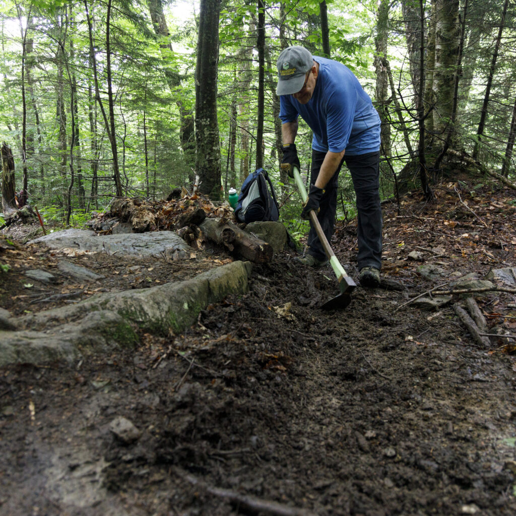 A volunteer uses a hand tool to scrape away soil and debris from part of the trail.